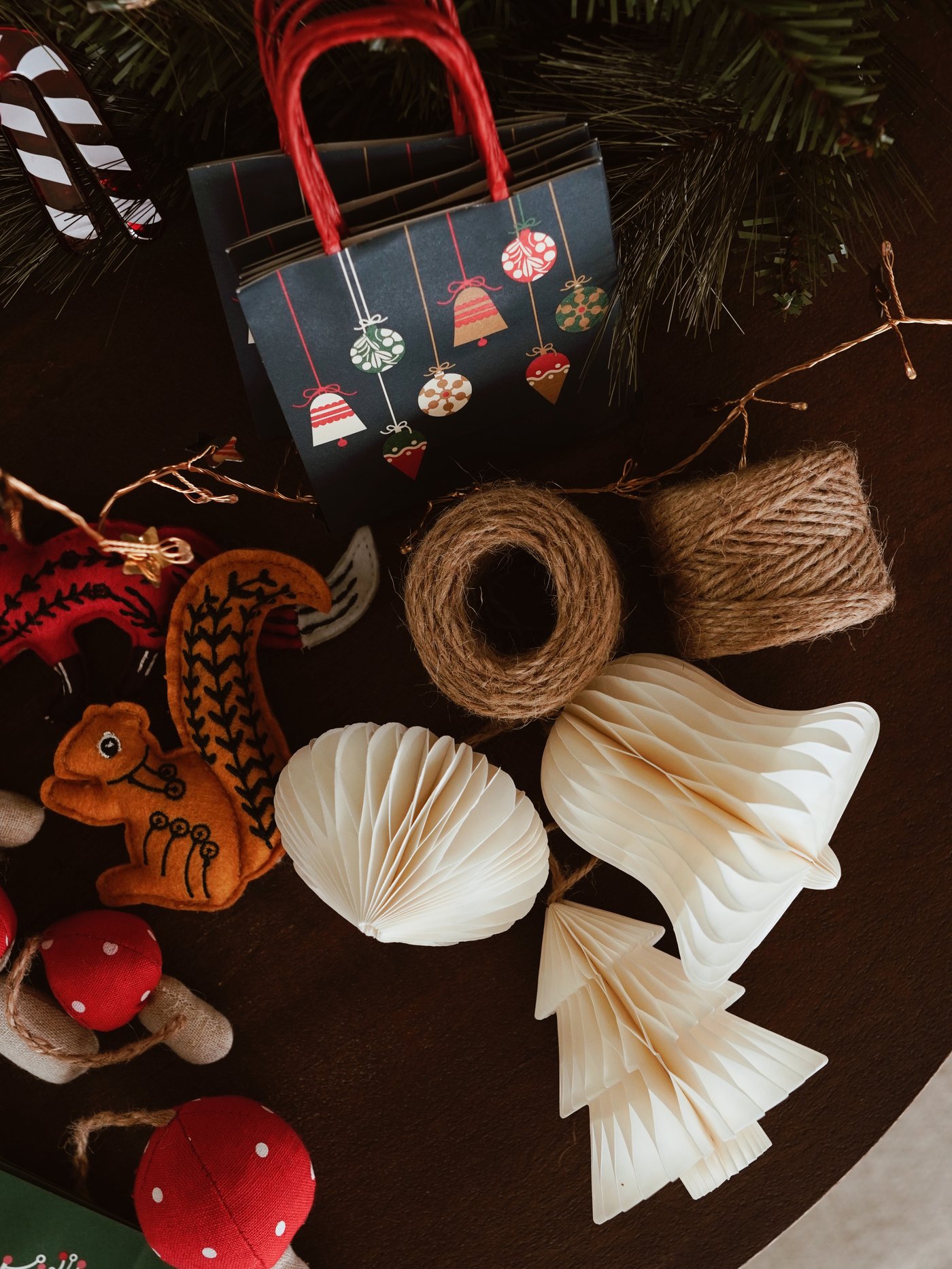 Una decorazione festiva per la tavola con ornamenti di carta, pigne, un rocchetto di filo e decorazioni in feltro accanto a un sacchetto regalo scuro con motivi festivi.