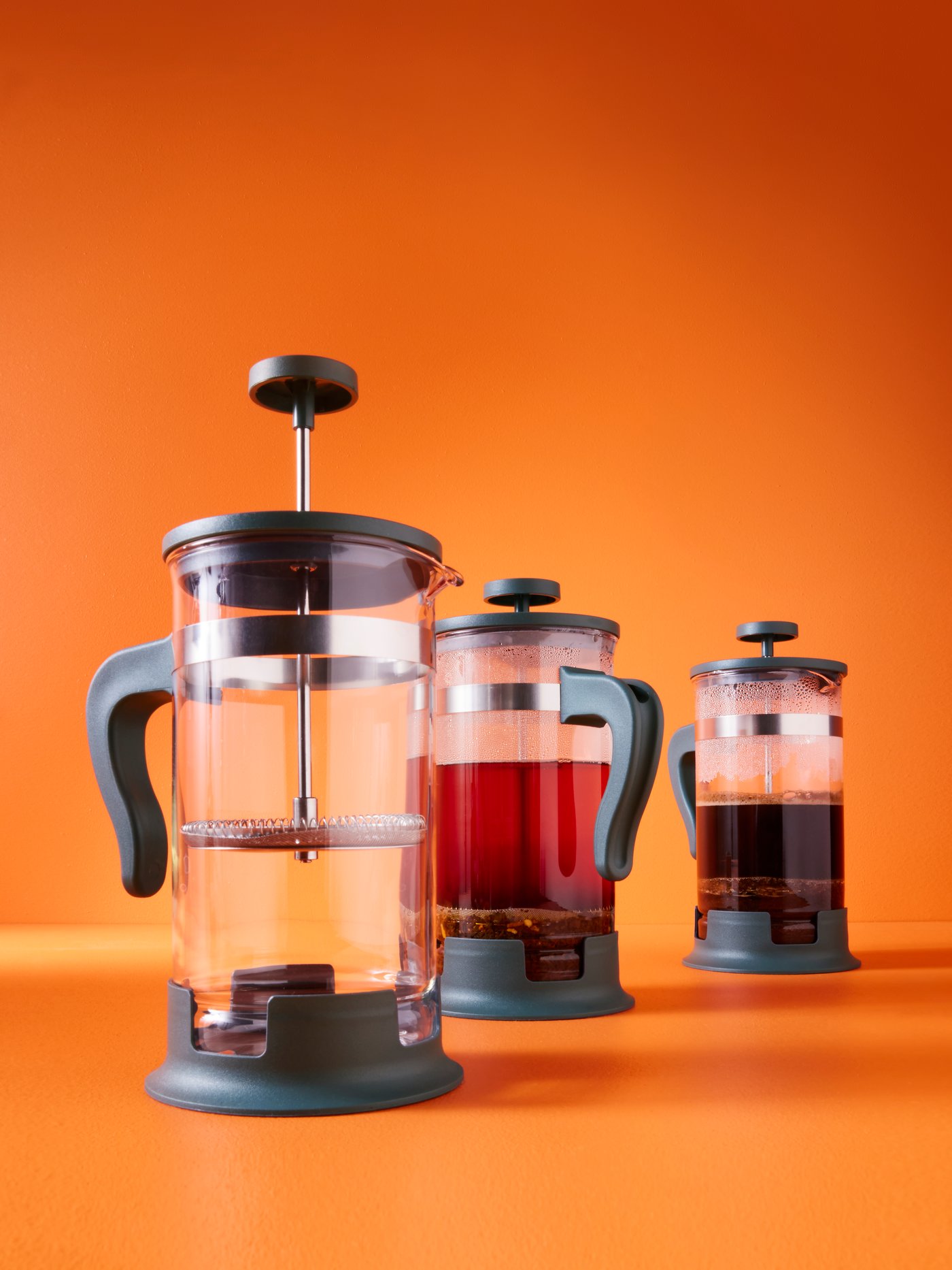 Three UPPHETTA coffee/tea makers, one containing coffee, the other containing tea, are placed on a bright orange surface.