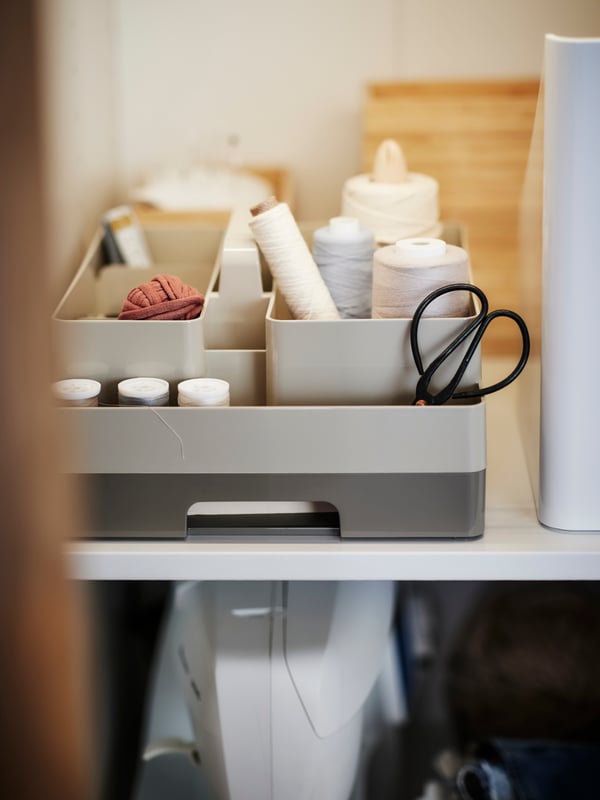 A grey-beige 2-piece KUGGIS desk organiser on a shelf filled with sewing thread and accessories in various colours.