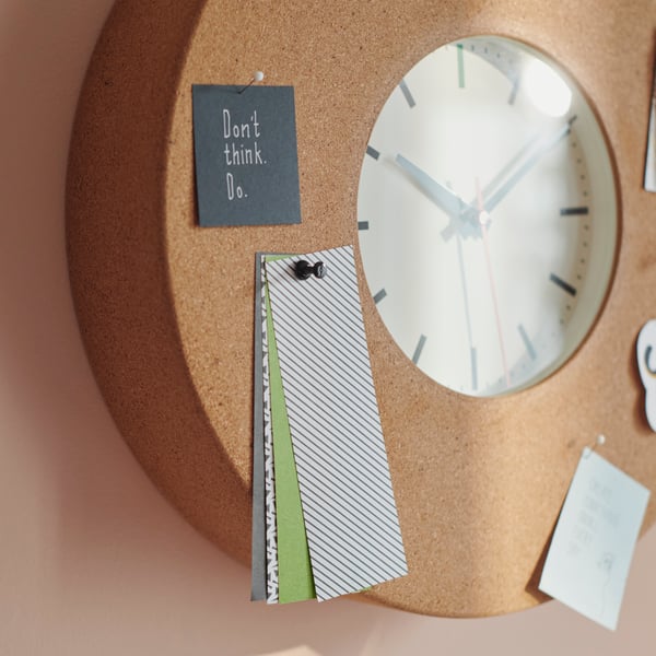 A SVARTGÖK wall clock, with various pieces of card pinned to its cork frame, is mounted on a wall.