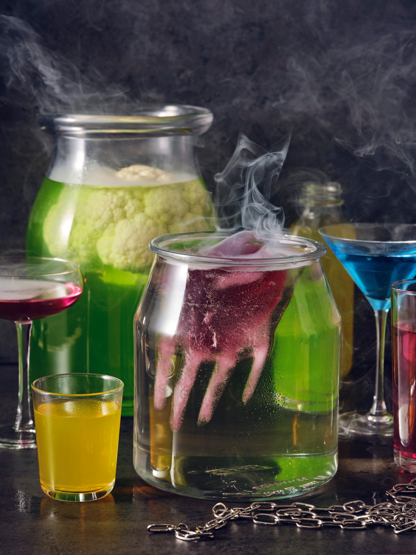 Een mix van doorzichtige glazen en potten, waaronder een IKEA 365+ pot, bevat een rokerige, kleurrijke vloeistof, waardoor een griezelige sfeer ontstaat.