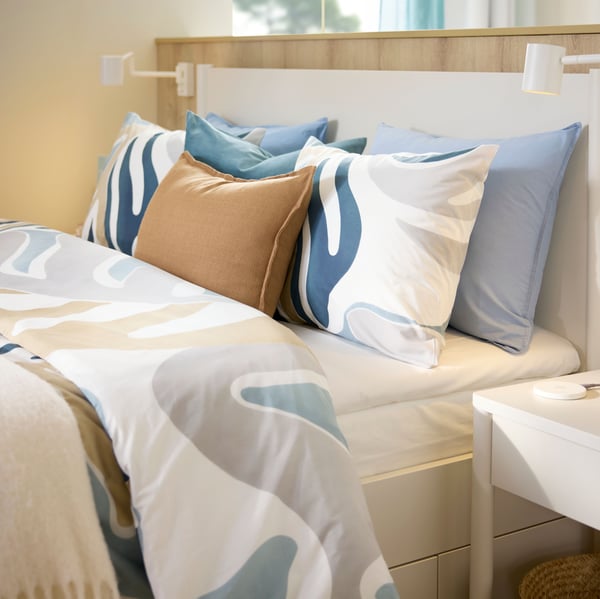 A hotel bed by a low wooden dividing wall is made with MYGGLASVINGE duvet cover and pillowcases that has a big leafy pattern.
