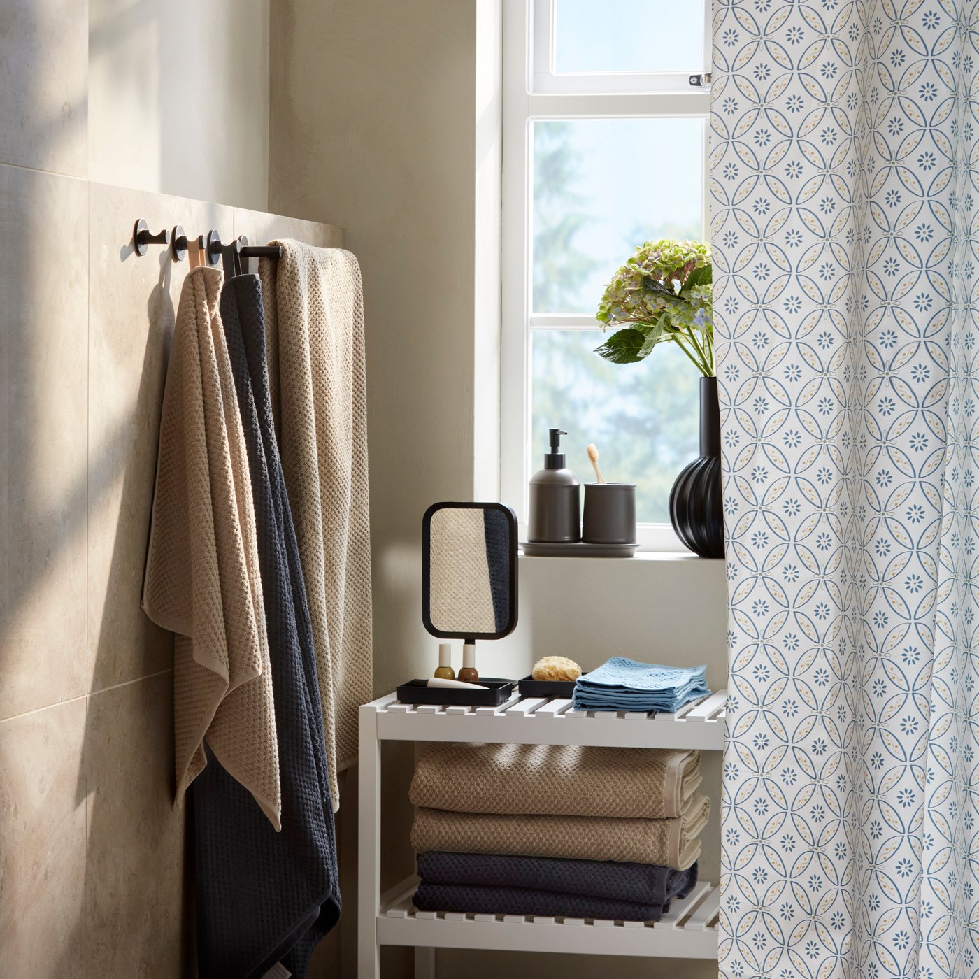 A bathroom corner has a black-framed LINDBYN table mirror on a bench by black trays, folded towels and a shower curtain.