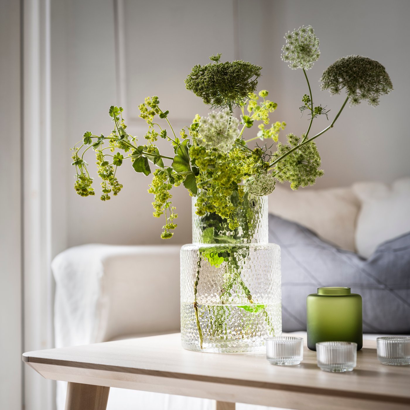 Kirkaslasinen KONSTFULL-maljakko, jossa värikkäitä kukkia olohuoneen sohvapöydällä.