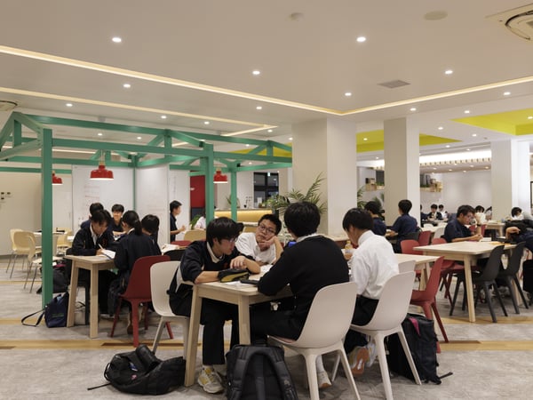 緑の柱と赤い照明が印象的な室内で、複数の人がテーブルに座って作業している様子