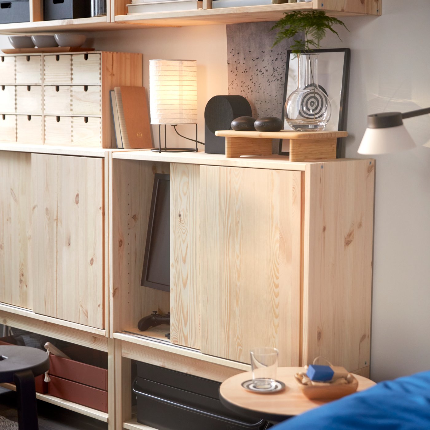 Two IVAR cabinets in pine with sliding doors displaying decorative objects and a lamp on top, placed next to a side table.
