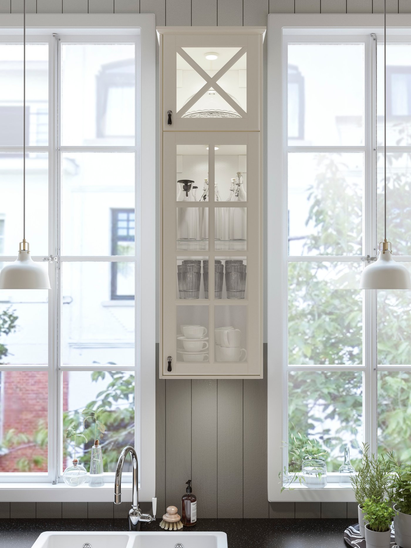 Kabinet dapur berwarna putih dengan pintu kaca vitrin BODBYN IKEA di antara dua tingkap sempit dengan dua lampu pendan berwarna putih.