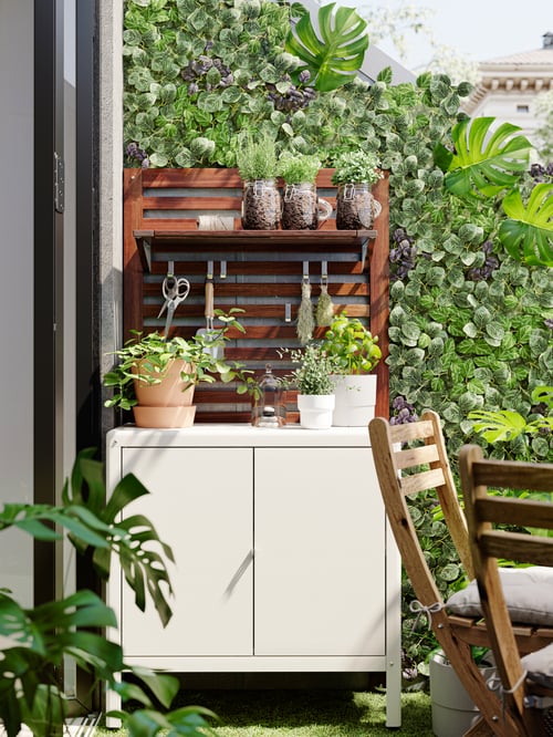 Zonă de exterior cu dulap bej pentru exterior, masă cu două scaune pliante, plante artificiale, ghivece de plante, raft, panou de perete.