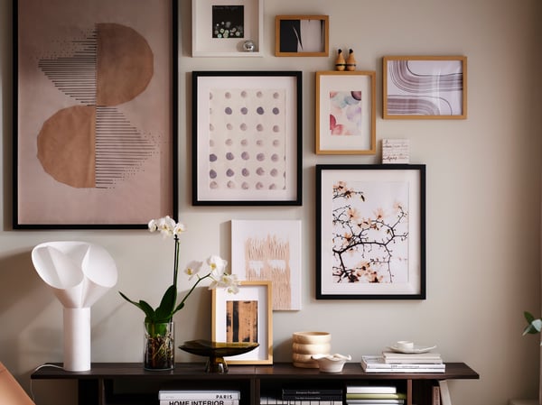 Un mur dans un salon lumineux, orné d’un groupe d’œuvres d’art présentées dans des cadres RÖDALM, au-dessus de tables d’appoint HOLMERUD ornées d’orchidées et d’objets déco.