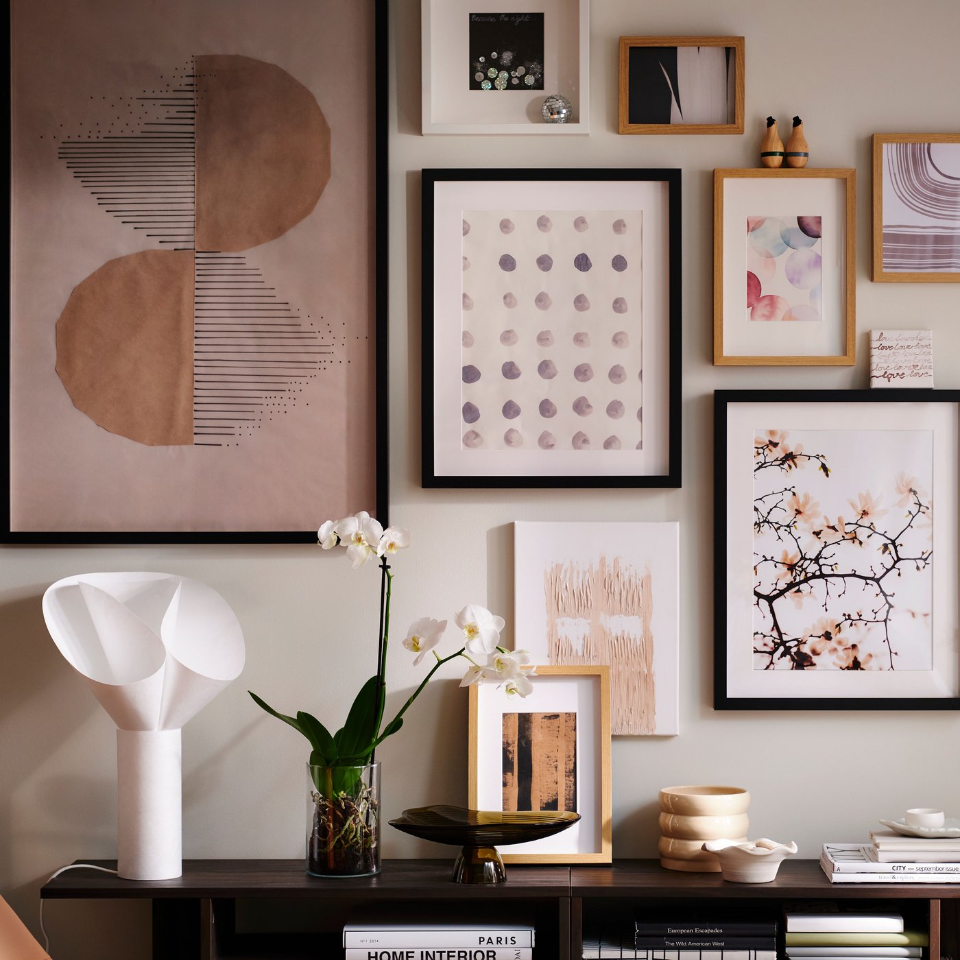 A wall in a light living room with an art cluster in various RÖDALM frames above HOLMERUD side tables with orchids and deco.