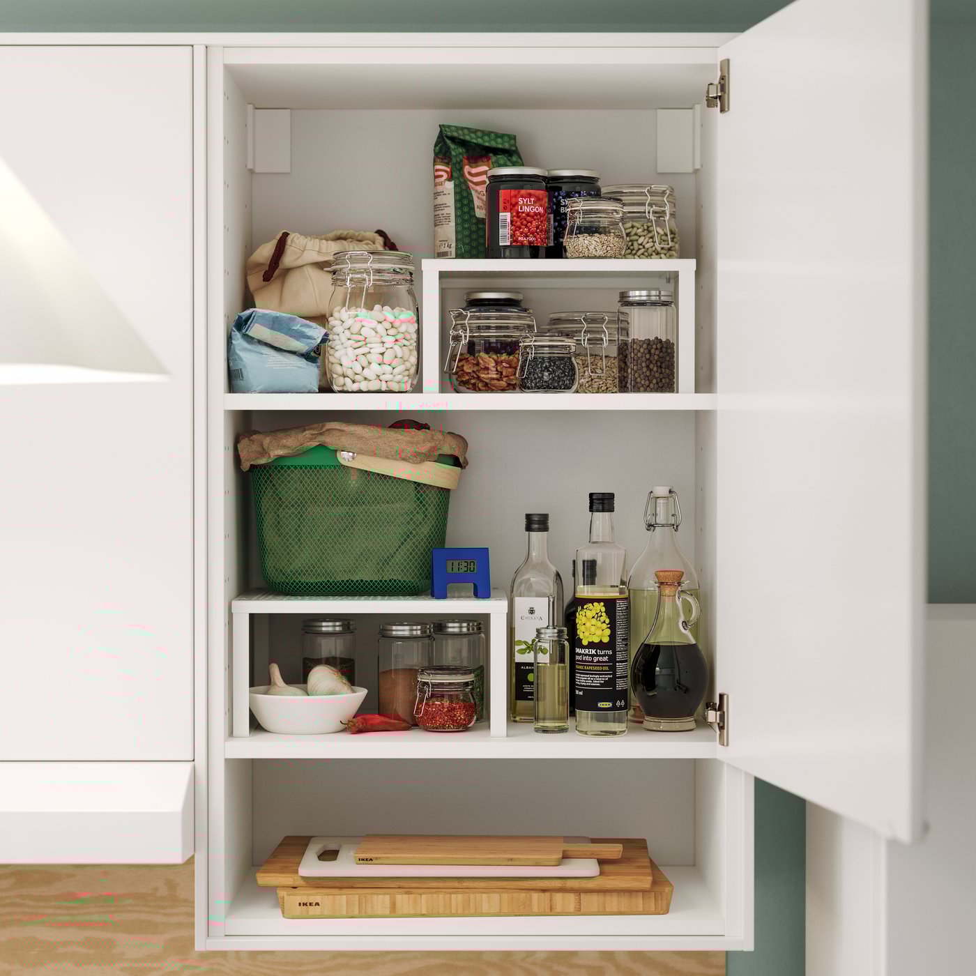 Balta VARIERA lentynos įdėklas laiko stiklainius ir butelius, tvarkingai sudėliotus modernioje virtuvės spintelėje kartu su maisto produktais.