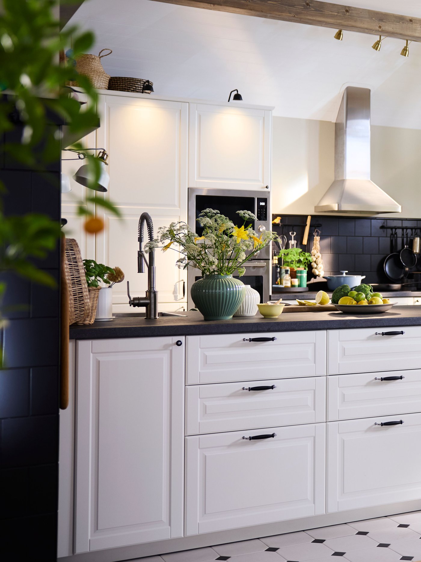 Cuisine lumineuse et confortable avec des armoires blanches, des plans de travail noirs et un îlot central. L'espace est décoré de plantes vertes, de produits frais et baigné de lumière naturelle provenant de grandes fenêtres. Une petite table à manger avec des chaises noires se trouve à droite, sous deux suspensions blanches.