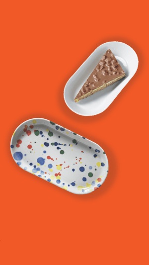 Farfurie albă ovală cu o felie de prăjitură cu ciocolată și blat crocant, alături de o tavă ovală cu model stropit colorat, pe fundal portocaliu intens.