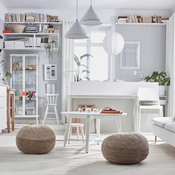 Un soggiorno molto luminoso in stile scandinavo moderno sui toni del bianco con due pouf SANDARED, un tavolino KRAGSTA e sgabelli SKYRRE. Sulla parete di fondo, una vetrina MILSBO, mensole bianche con sopra libri, contenitori porta riviste e piante. Nella stanza ci sono tre lampade bianche a sospensione, di cui una REGNSKUR / SUNNEBY.
