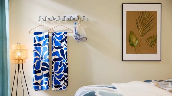 Wavy, dark grey-green JORDMÅNEN hooks hold beach towels and snorkel gear by artwork on a wall behind a neatly made hotel bed.
