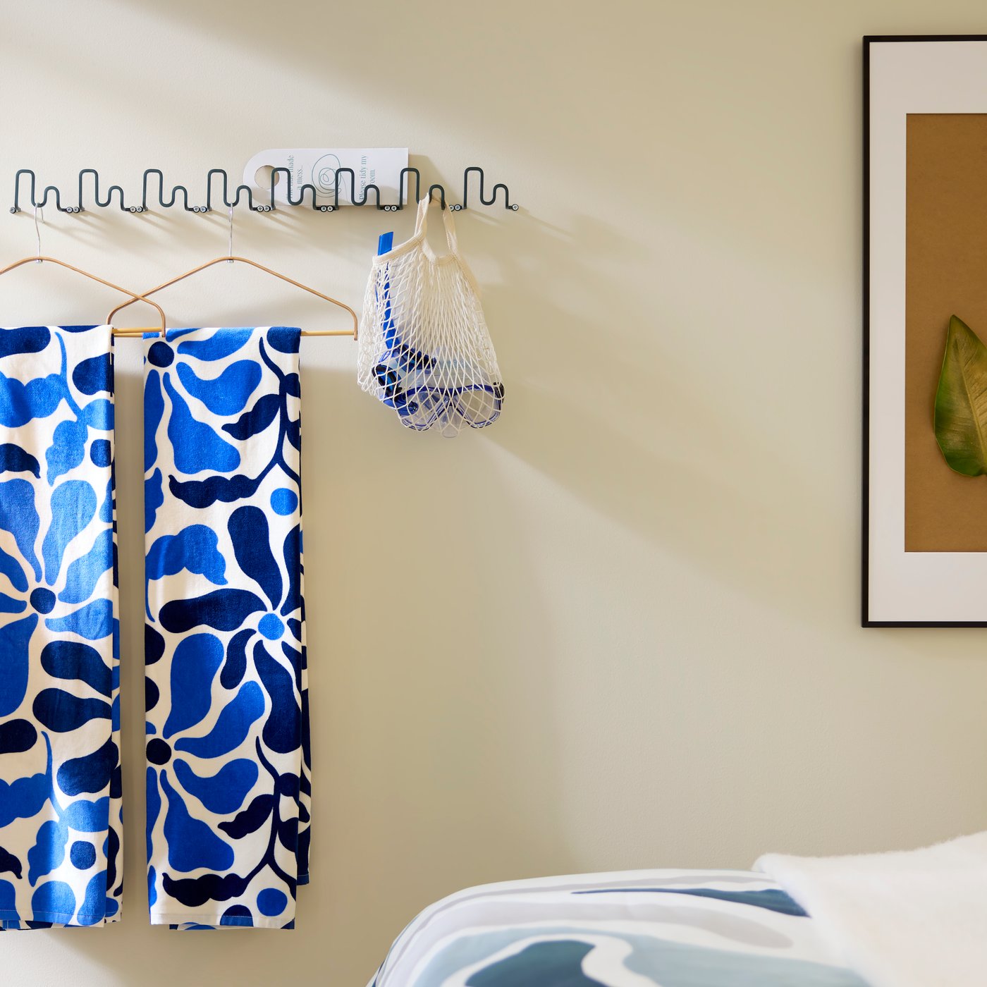 På bølgende, mørkegrå-grønne JORDMÅNEN kroge hænger strandhåndklæder og snorkeludstyr ved kunst på væggen bag den pænt redte hotelseng.