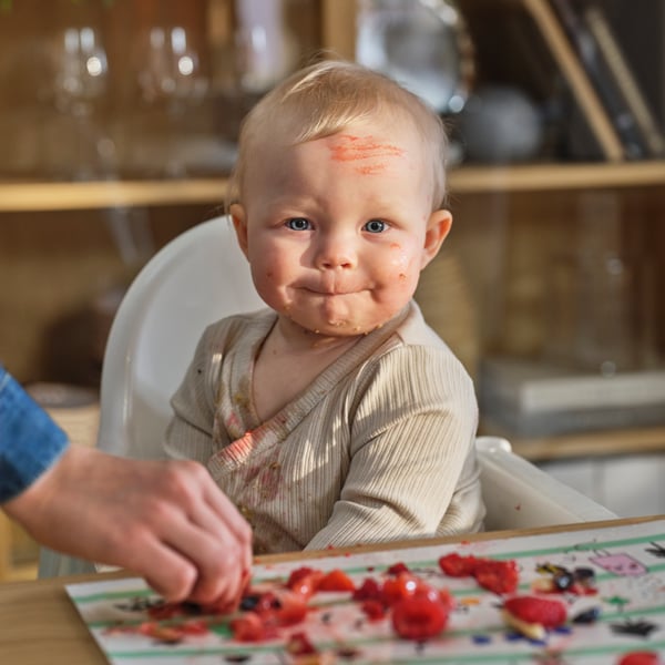A child sits in an ANTILOP highchair in front of a table with a GRÖNFINK place mat on it that’s covered in pieces of fruit.
