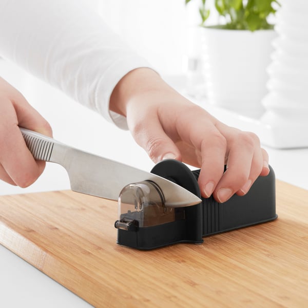 A pair of hands is holding a black ASPEKT knife sharpener on a wooden chopping board while sharpening an IKEA 365+ stainless steel knife.