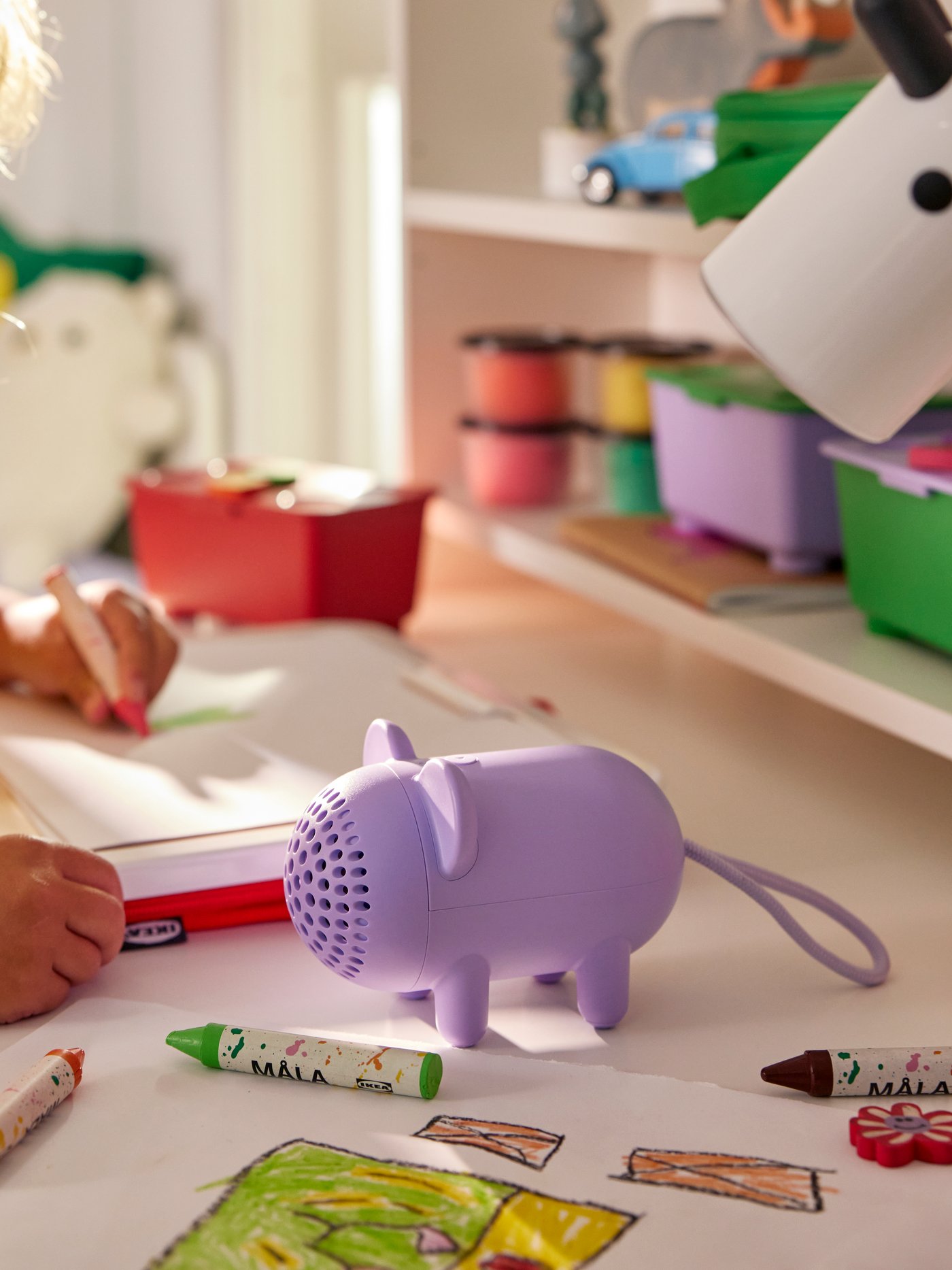 In a sunny kid’s room, a child at a desk draws a picture. A mouse-shaped GREJSIMOJS portable Bluetooth speaker is beside her.