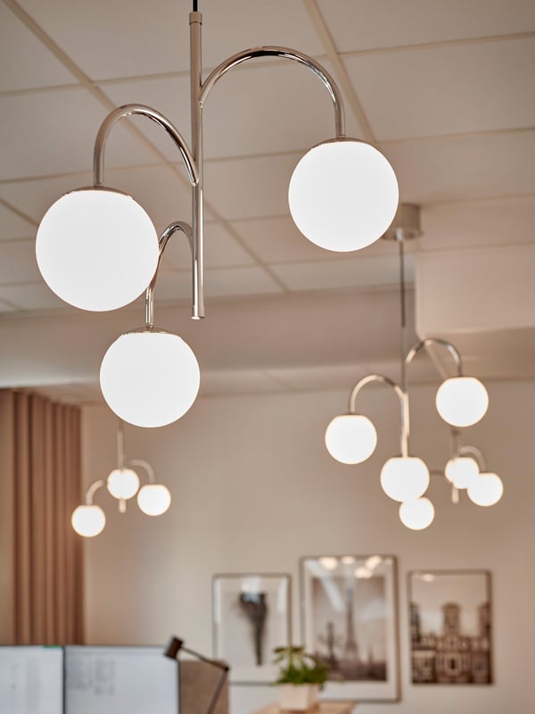 Interiör med flera SIMRISHAMN taklampor bestående av tre vita klotlampor på metallarmar, samt tavlor på väggen, växt på bord och kontorsmiljö i bakgrunden med skärmar och gardiner.