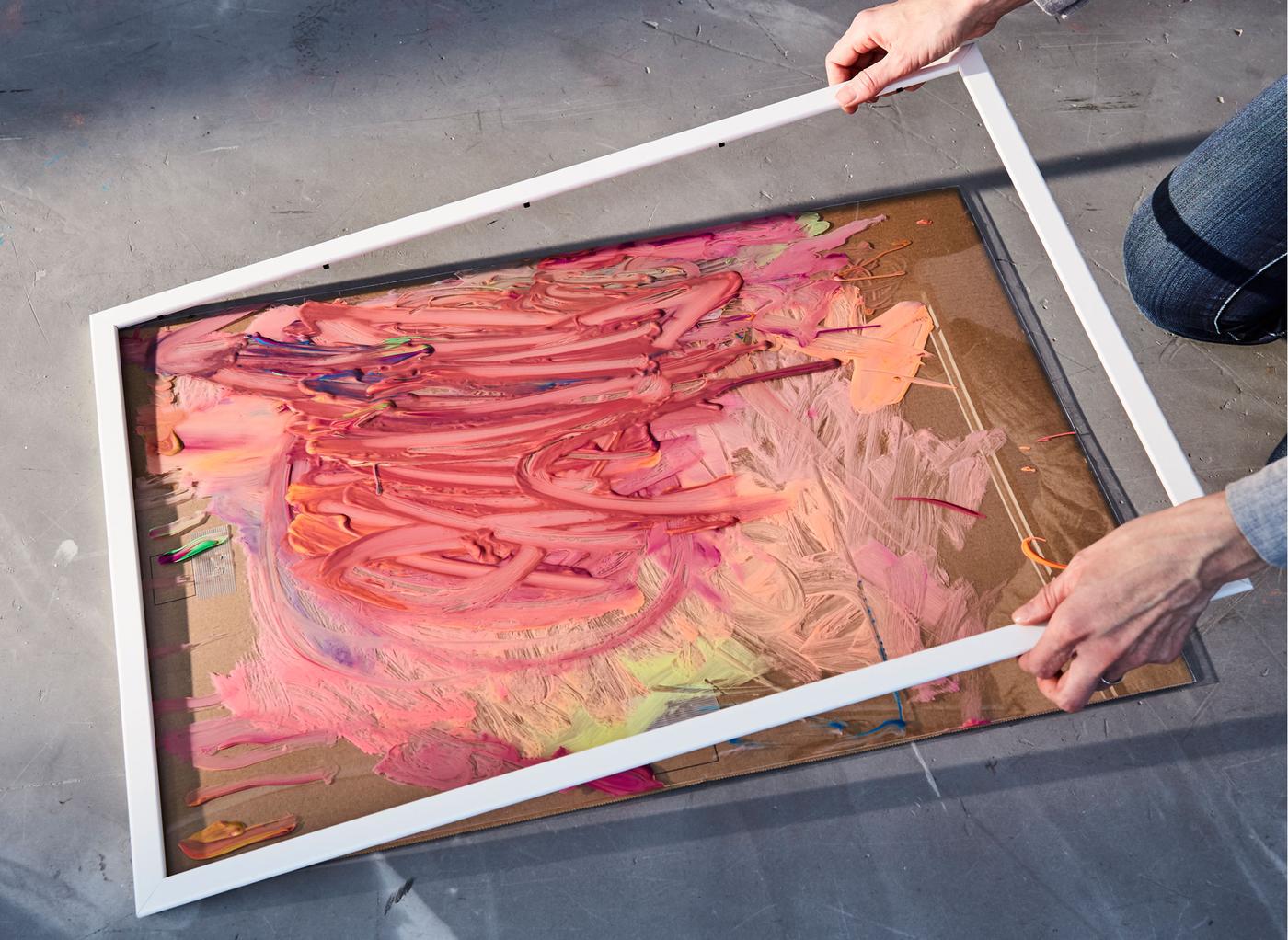 Zwei Kleinkinder drücken Farbe auf Pinsel und spielen mit Kreide. In einer Nahaufnahme ist ein Kindergemälde in einem FISKBO Rahmen in Weiss zu sehen.