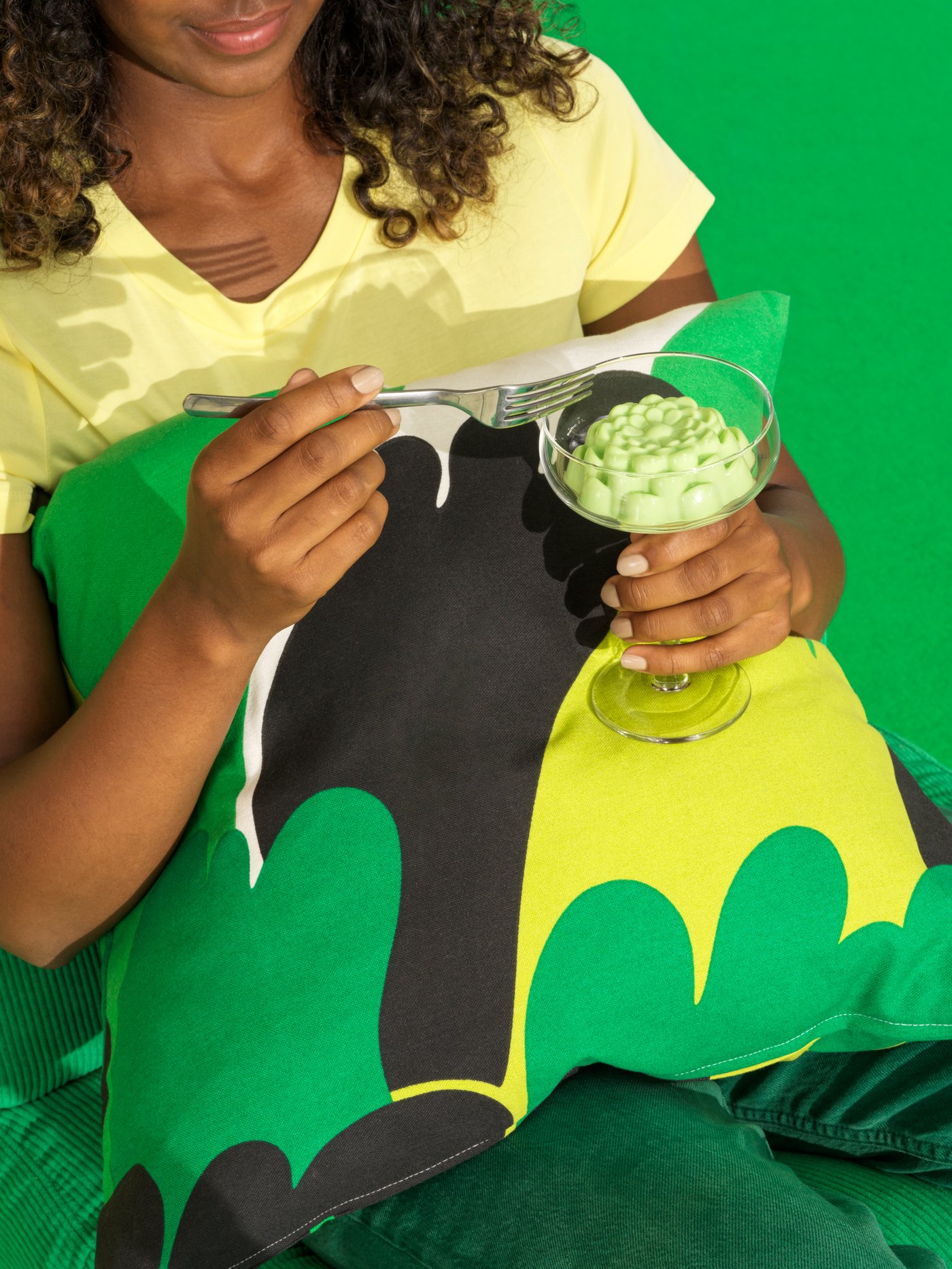A person in a green chair eats green pudding from a champagne coupe. In the lap is a cushion in an ÄNGSFIBBLA cushion cover.