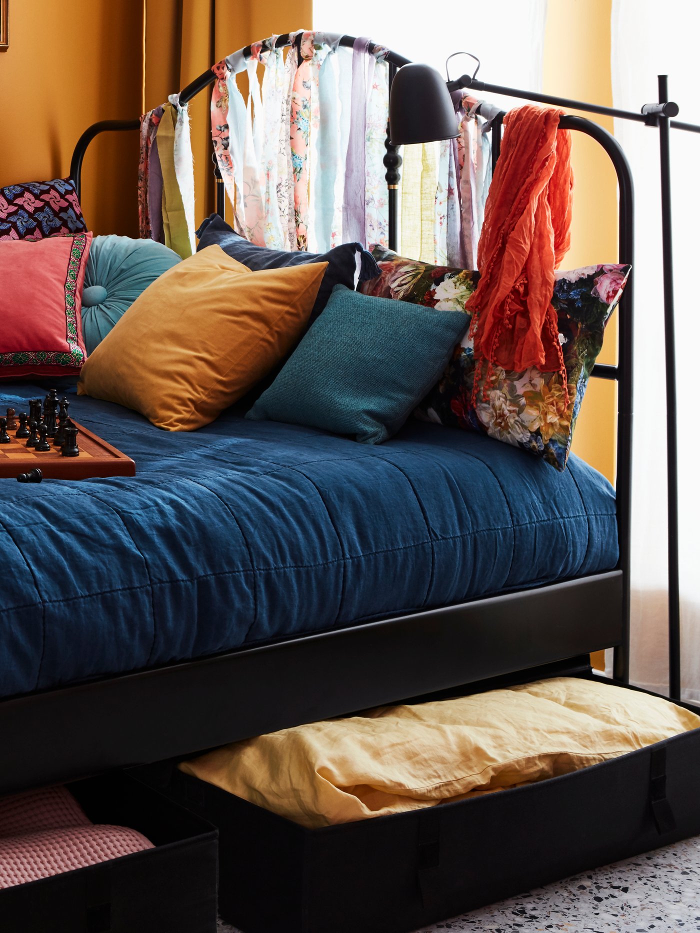 O cutie de depozitare sub pat LYCKSELE neagră, în care se află o lenjerie de pat de primăvară, sub un pat SAGSTUA cu lenjerie de pat colorată.