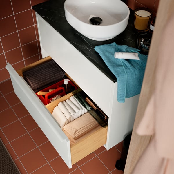 A white HAVBÄCK/VINDEÄLVEN washbasin cabinet with an open drawer showing organised items, topped with a folded blue towel.