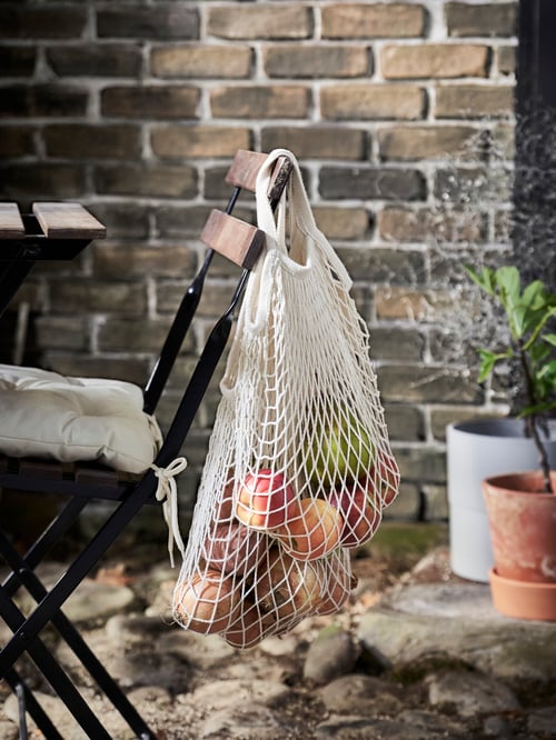 Outdoor area with two net bags with fruit, hung on the backrest of a chair, with cushion on seating area, and potted plants.