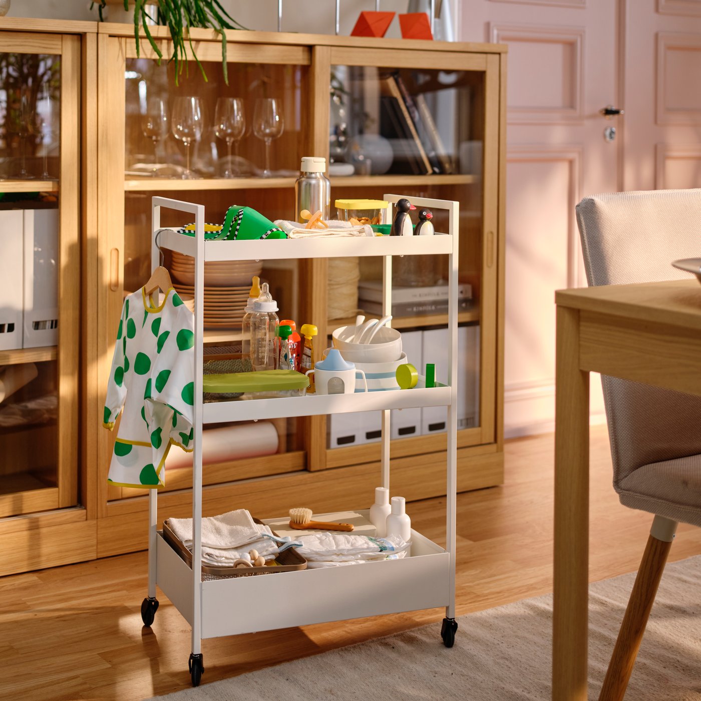 A white NISSAFORS trolley in a dining room holds baby feeding equipment including a bib and BÖRJA plates, bowls and spoons.