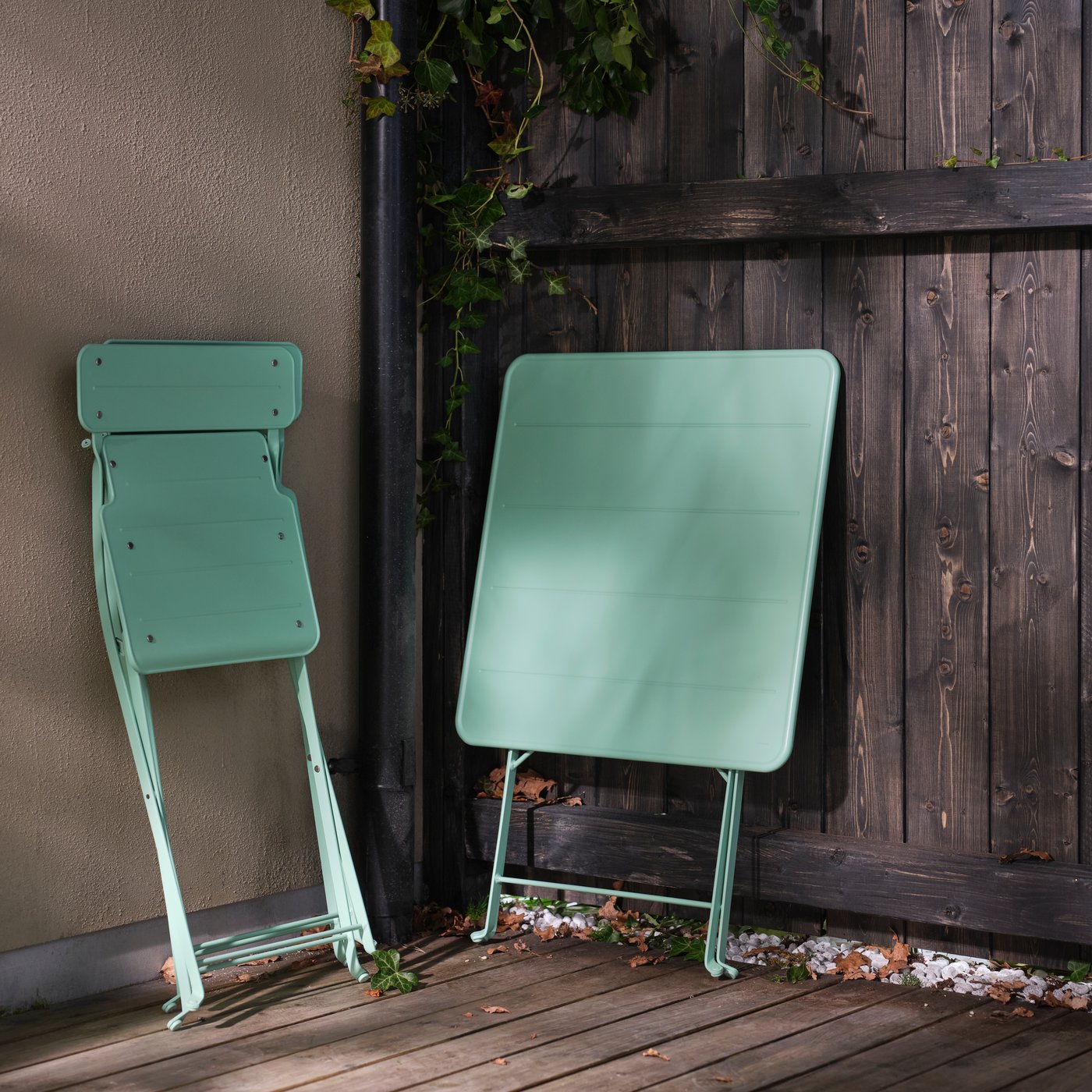 A folded SUNDSÖ table and a folded SUNDSÖ folding chair lean against a fence and a wall on a terrace.