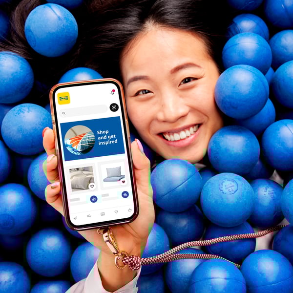 A smiling woman lies among blue balls, holding a smartphone that displays the IKEA app.