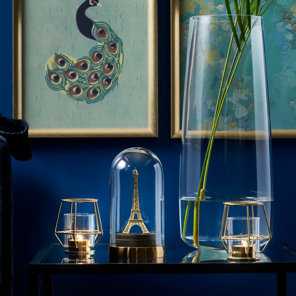 A clear glass BERÄKNA vase in the living room, next to a BEGÅVNING glass dome covering an Eiffel Tower ornament. Next to it is a PÄRLBAND tealight holder. On top of it is a SILVERHÖJDEN frame filled with artwork.