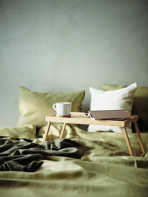 Bedroom with bed with duvet, linen, blanket, three cushion covers, bamboo bed tray, white mug, notebook with brown cover.