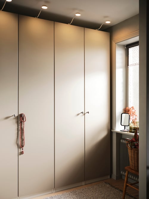 A beige PAX/REINSVOLL wardrobe combination beside a window with white YTBERG cabinet lighting along the top.