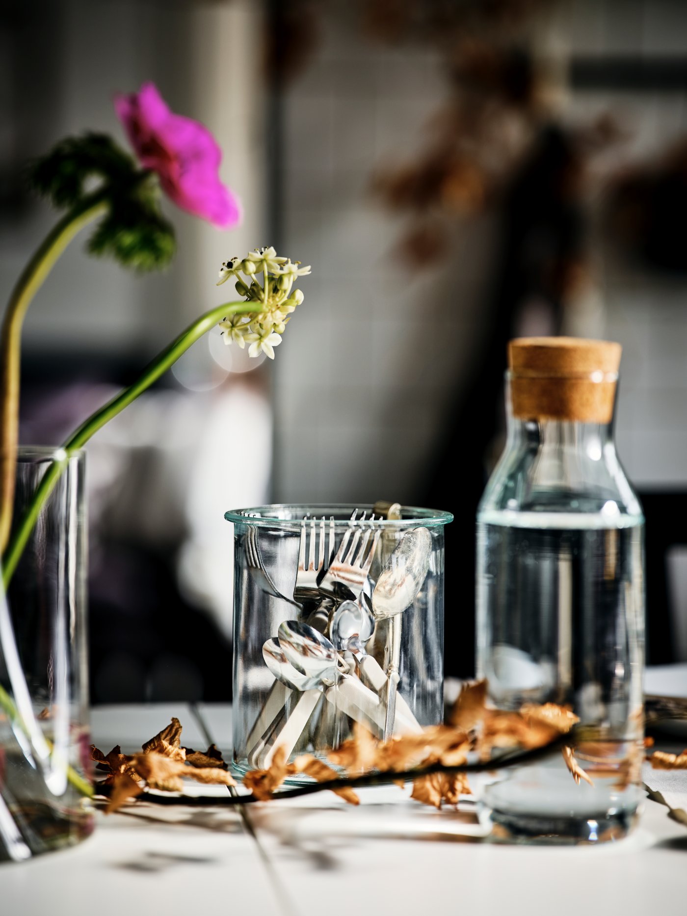 A tabletop with an IKEA 365+ clear-glass carafe with a cork, cutlery gathered in a glass jar, and two flowers in a vase.