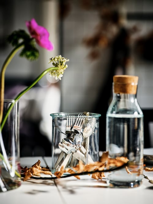 O masă cu o carafă din sticlă transparentă IKEA 365+ cu dop de plută, mai multe tacâmuri într-un borcan de sticlă și două flori într-o vază.