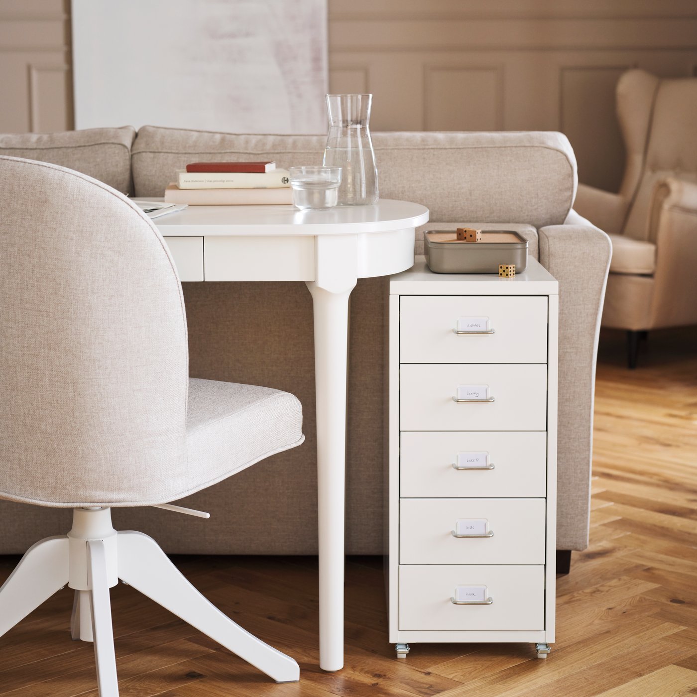 Un fauteuil pivotant ROSENTORP et une commode blanche HELMER à roulettes sont placés devant un bureau blanc ROSENTORP, derrière un canapé.
