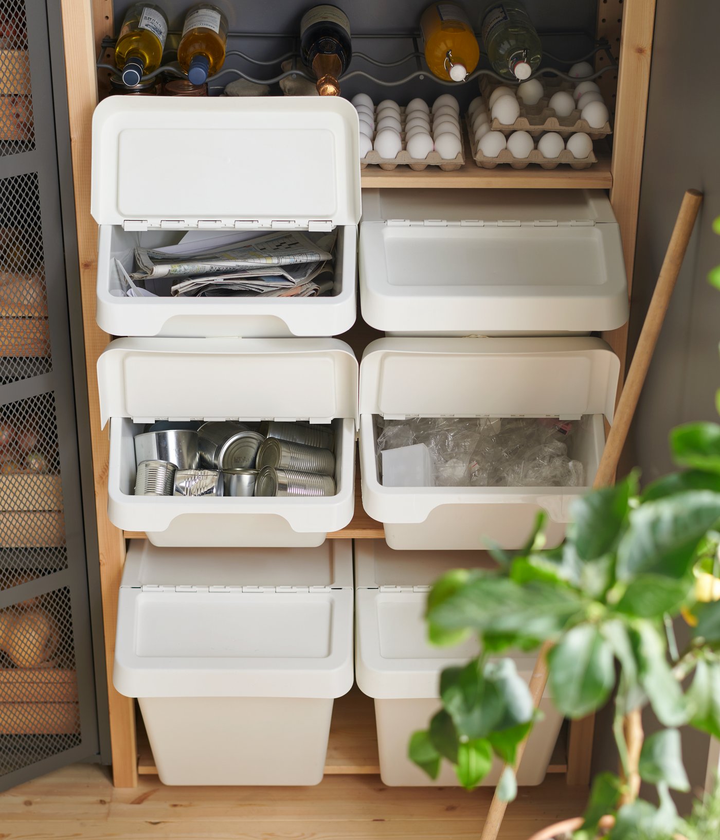 White SORTERA waste sorting bins with lids stacked on a wooden shelf containing recyclable paper, plastic and metal items.