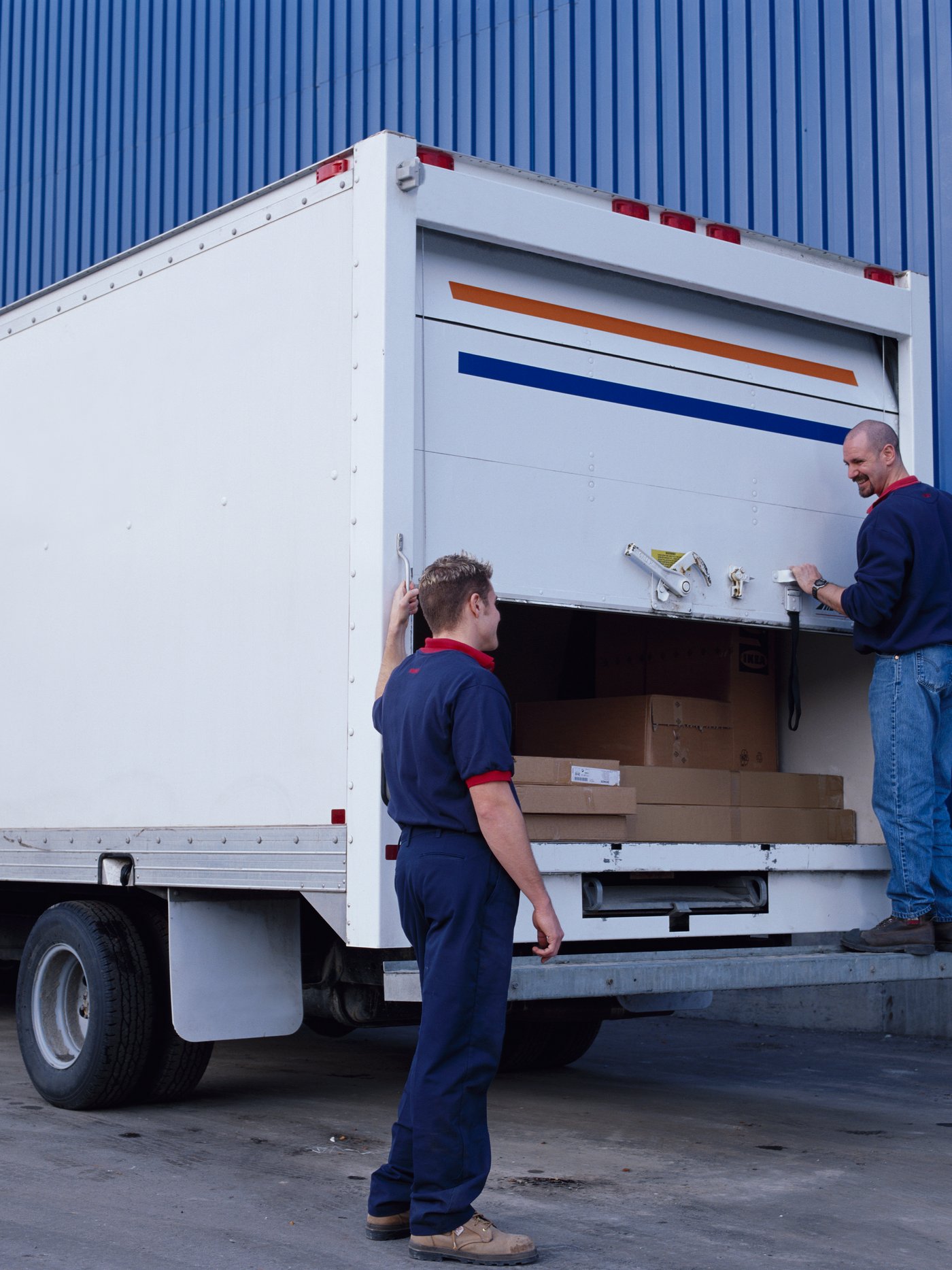 L’image montre deux livreurs en train de charger ou décharger un camion blanc garé devant un bâtiment bleu, typique d’un entrepôt IKEA. On aperçoit à l’intérieur du véhicule plusieurs colis plats et cartons, caractéristiques des emballages de meubles. La scène illustre la logistique et l’organisation derrière la livraison, dans un cadre professionnel et structuré.