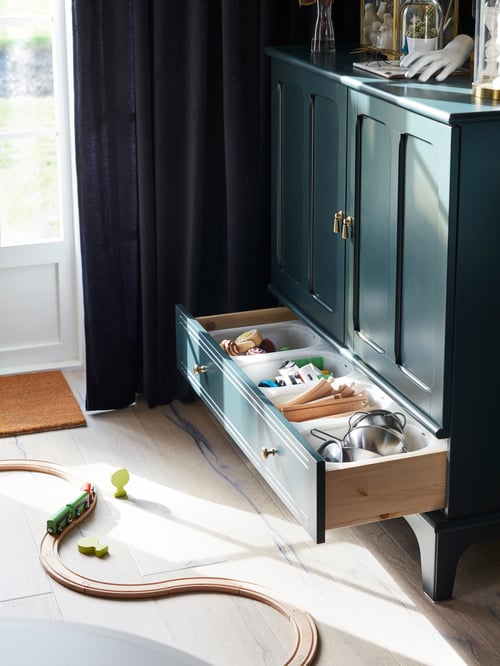 Modern room with dark blue-green cabinet next to windows with room darkening curtains, orange door mat and basic train set.