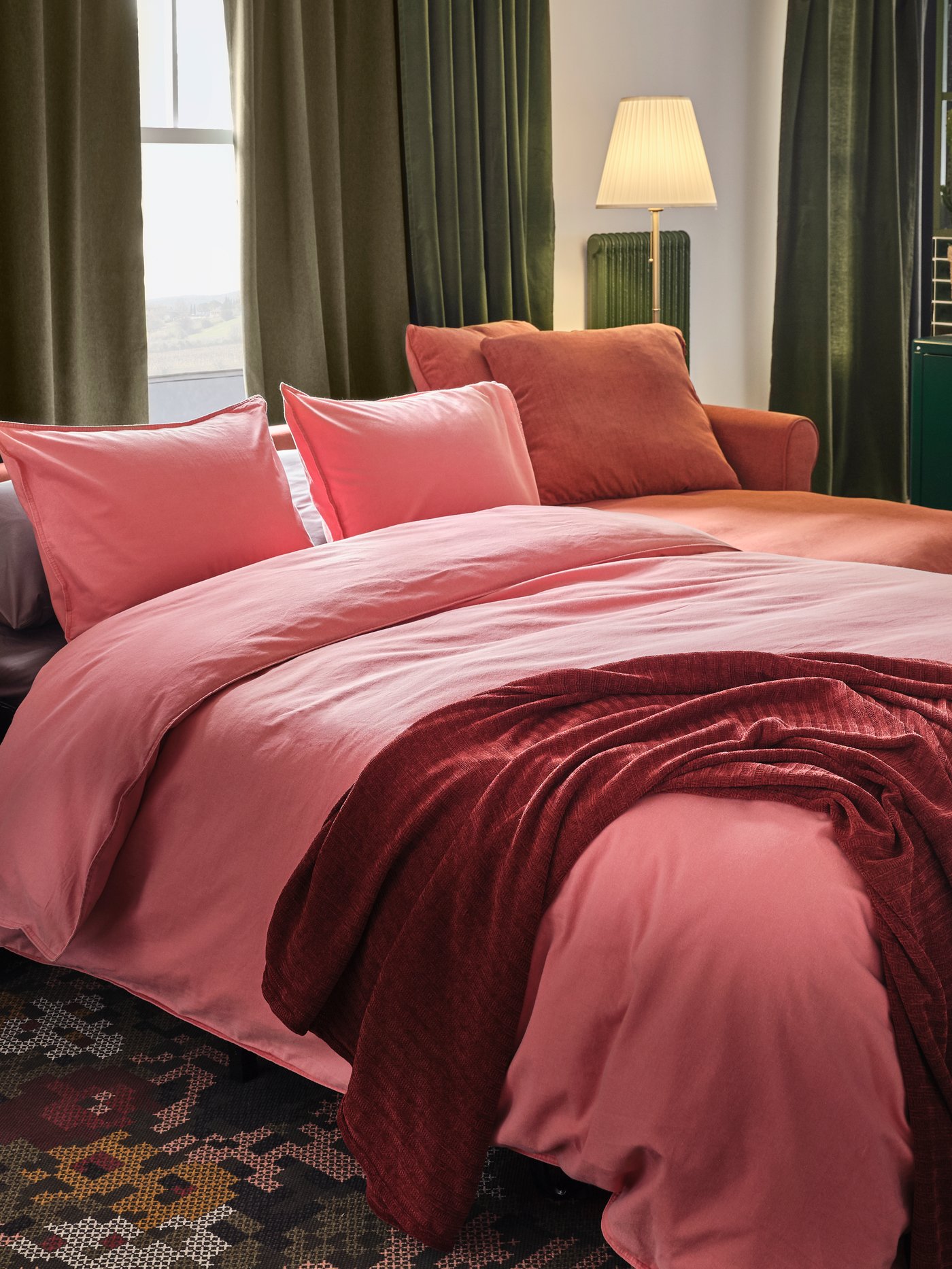 A light red 3-seat sofa-bed made as a bed with light brown-red bed textiles. Green curtains cover two windows behind.