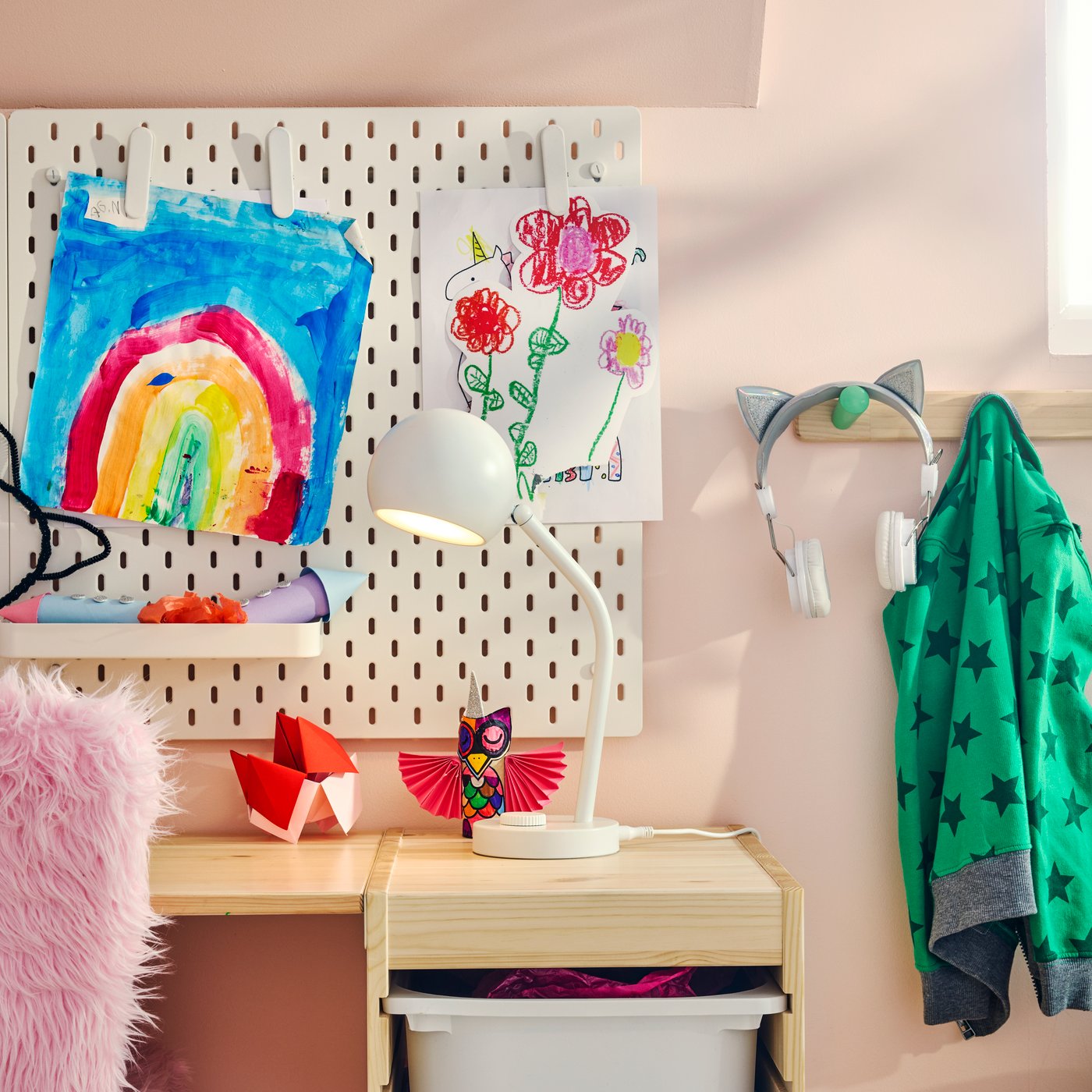 A pink children’s room with a white KORVSNÖRE lamp on a pine desk, beside a pegboard wall displaying children’s artwork.