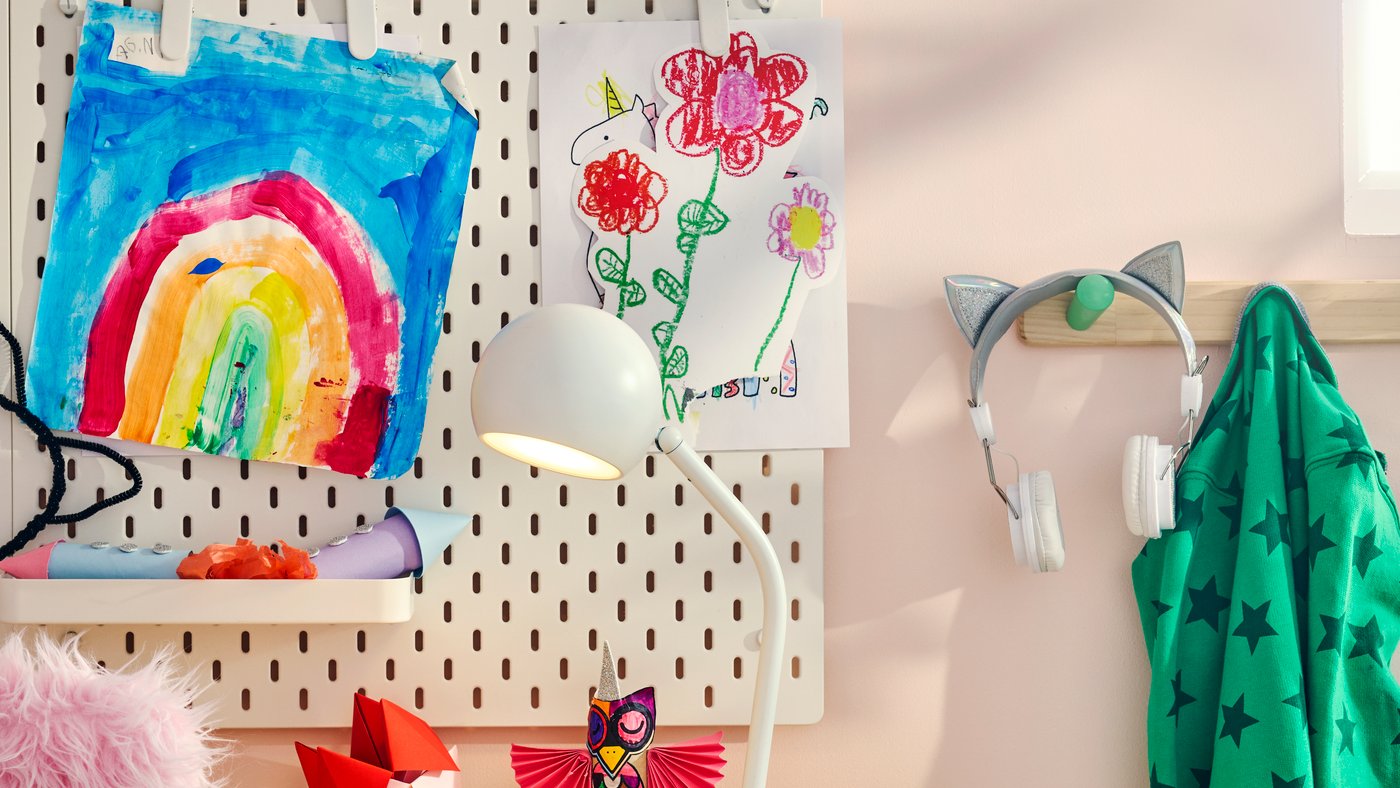 Ett rosa barnrum med en vit KORVSNÖRE-lampa på ett skrivbord i furu, bredvid en förvaringstavla som visar upp barnens konstverk.