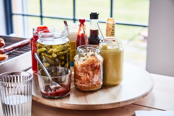 Platou rotund din lemn cu borcane și sticle de condimente: murături, varză murată, muștar, ketchup, sos de soia și alte sosuri. În prim-plan se află un pahar gol.