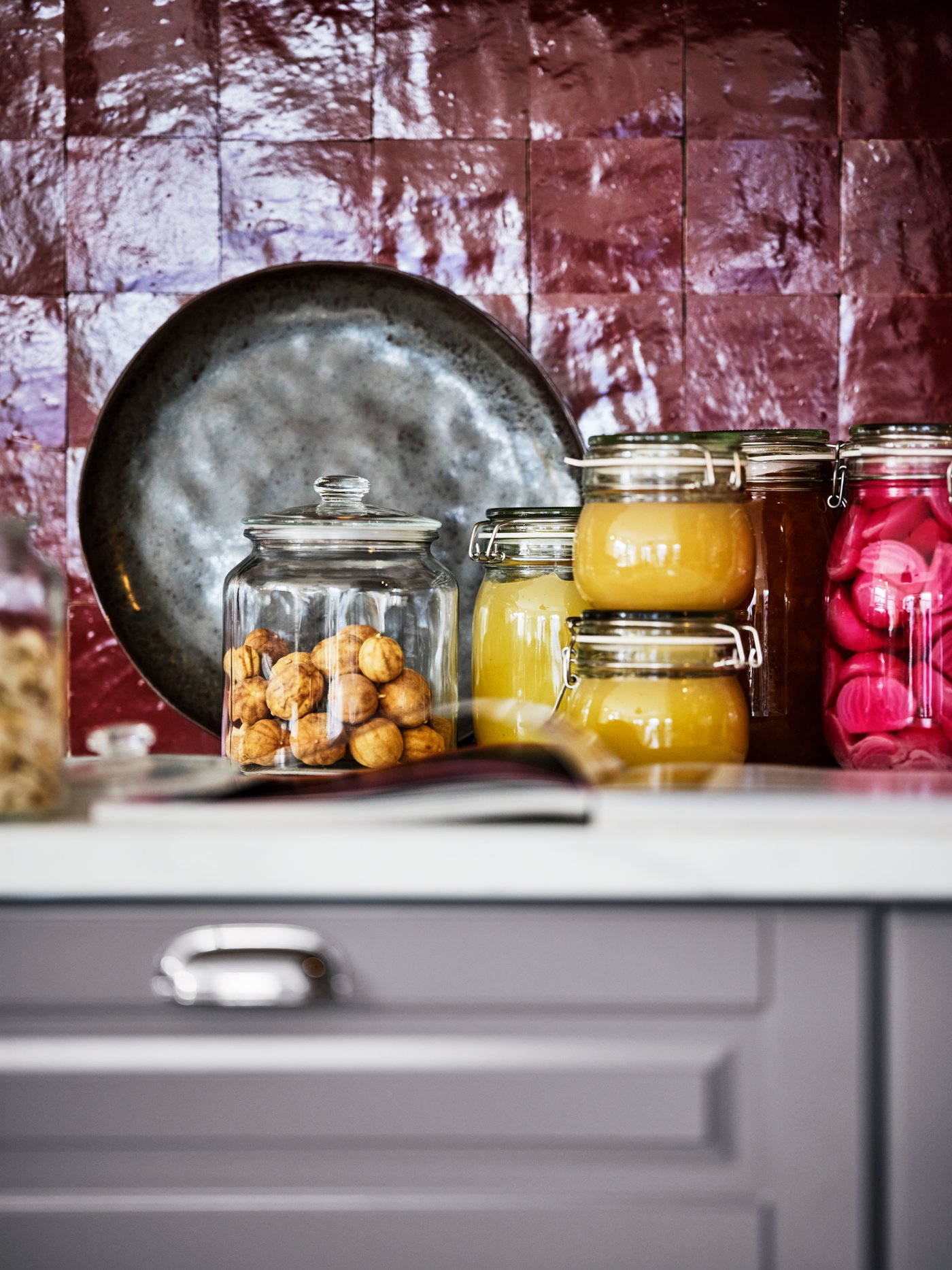 Un mobile con sopra contenitori con coperchio VARDAGEN e barattoli KORKEN con all'interno alimenti. Dietro, uno stampo appoggiato ad una parete con piastrelle rosso scuro.