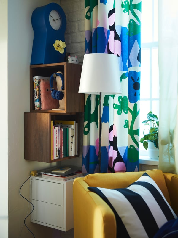 A yellow EKERÖ armchair placed next to a corner where three EKET cabinets have been mounted vertically, holding mainly books.