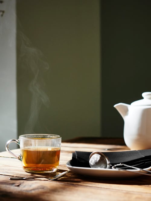 Dining area with wooden dining table, glass mug with tea, tea infuser, white teapot, black table napkin, teaspoon, plate. 