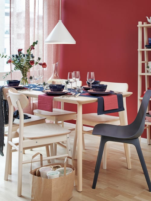 Dining area with red walls, dining table, chairs, napkins, plates, bowls, glasses, carafe, vase with flowers, pendant lamp.