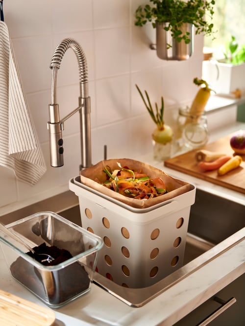 Kitchen sink with mixer tap with hand spray and white plastic bin with ventilation holes filled with vegetable scraps.